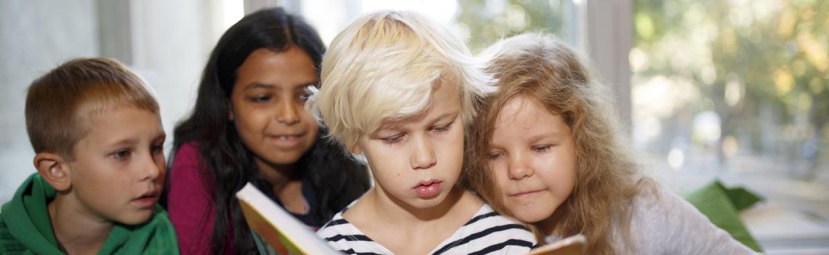 Etualalla noin 8-vuotias poika lukee kirjaa. Pojan takana samaa kirjaa kurkkii kaksi samanikäistä tyttöä.