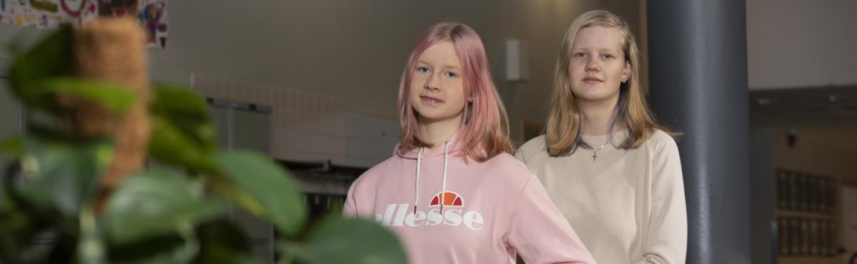 Kaksi yläkouluikäistä tyttöä seisoo rinnakkain kaiteeseen nojaten ja katsoo kameraan. 