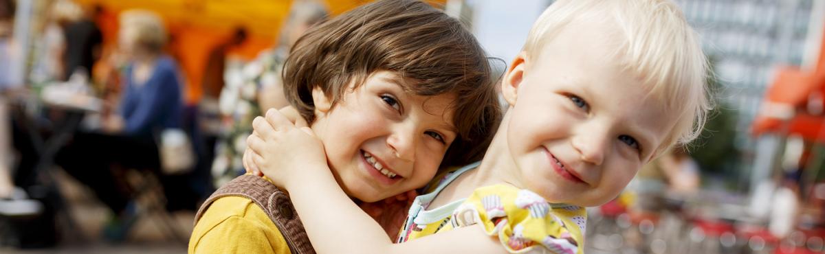 Leikki-ikäinen tyttö halaa tiukasti samanikäistä poikaa keskellä kesäistä torimiljöötä. 