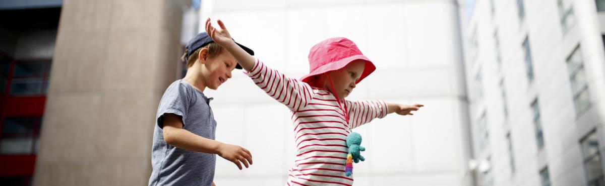 Noin 7-vuotias poika ja tyttö tasapainoilevat pihalla massiivisten kerrostalojen keskellä. Tyttö on levittänyt kätensä, poika tulee perässä. 