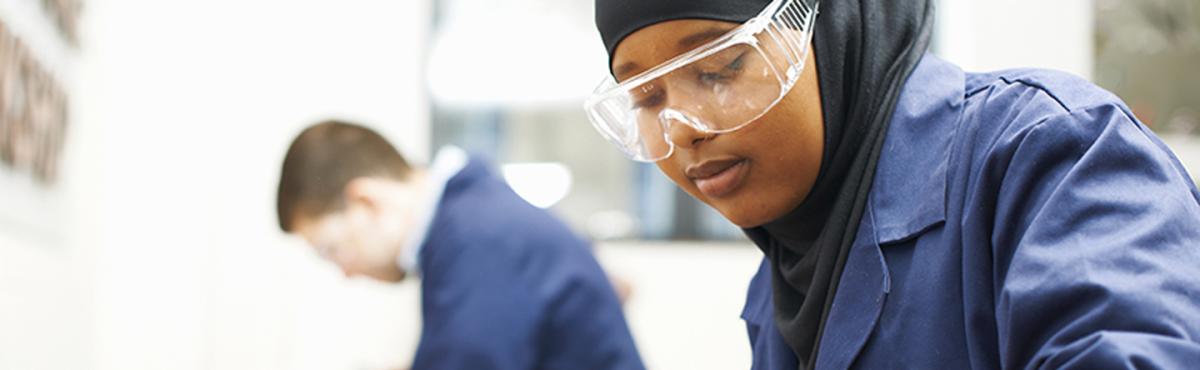 Suojalaseihin ja -takkiin sonnustautunut huivipäinen, teini-ikäinen tyttö katselee keskittyneesti työhönsä luokassa. 