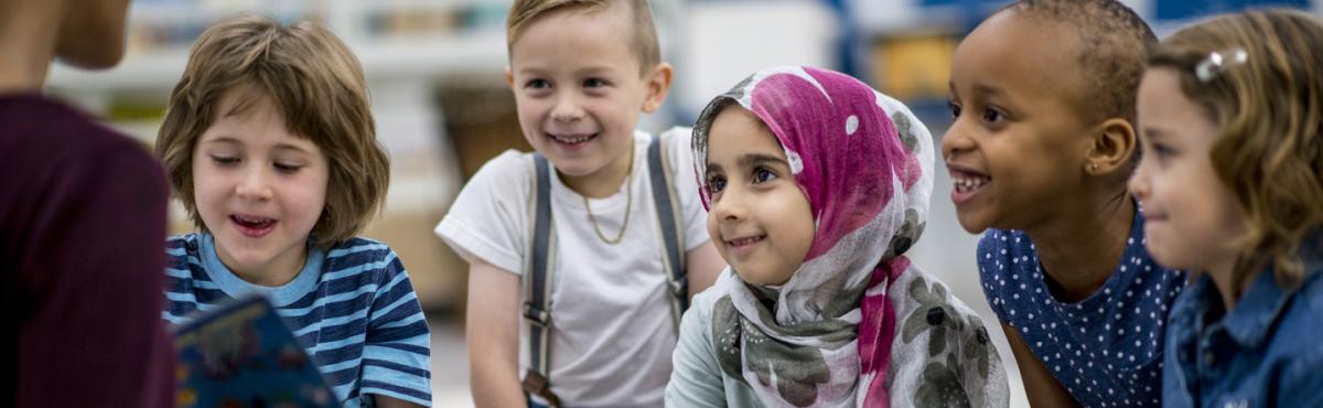 Viiden esikouluikäisen lapsen ryhmä istuu katselemassa kirjan aukeamaa, jota opettaja näyttää. 