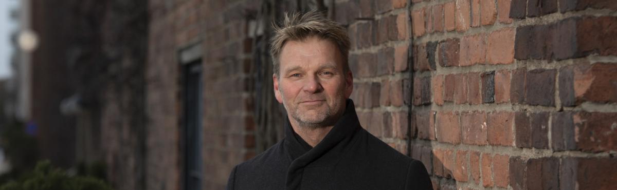 Mies katsoo kameraan, taustalla näkyy tiiliseinää. 