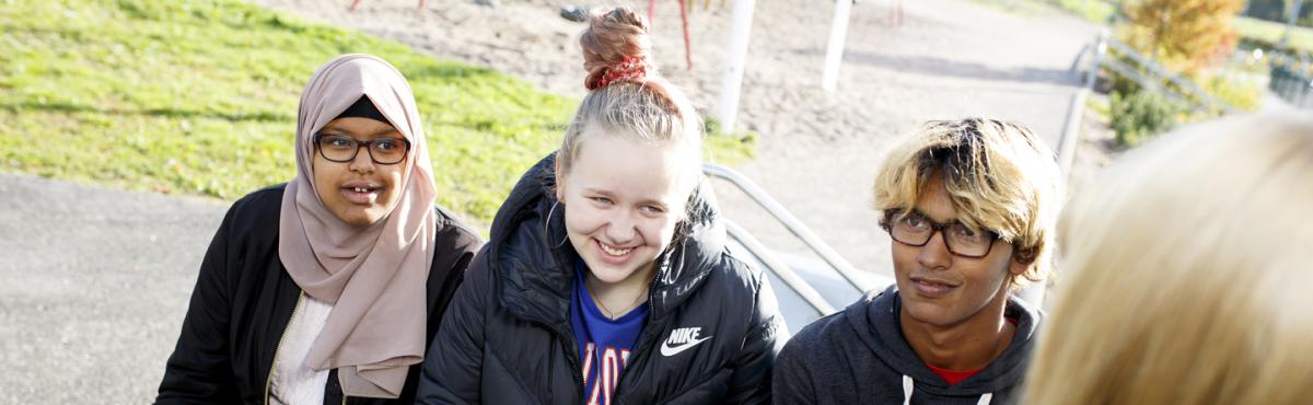 Kaksi teini-ikäistä tyttöä ja yksi poika istuvat koulun pihalla. 