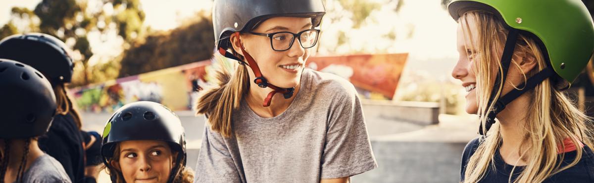 Kaksi teini-ikäistä tyttöä juttelee skeittipuistossa kypärät päässä. Taustalla näkyy nuorempi tyttö, jolla on myös kypärä päässä.