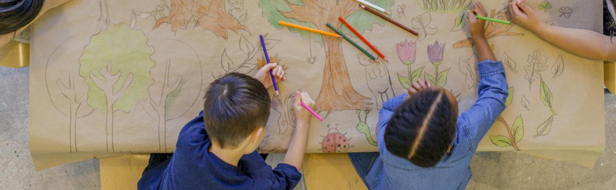 Ylhäältä päin kuvatussa kuvassa kaksi lasta piirtää värikynillä isolla ruskealle paperille puita, kukkia ja perhosia. 