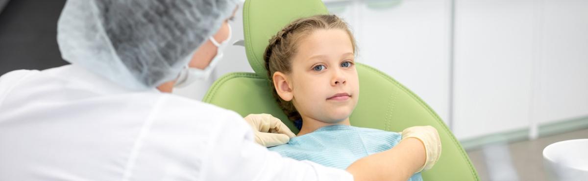Lapsi makaa vihreässä hammaslääkärin tuolissa ja etualalla valkoiseen takkiin ja maskiin pukeutunut henkilö asettelee hänelle suojaessua. 