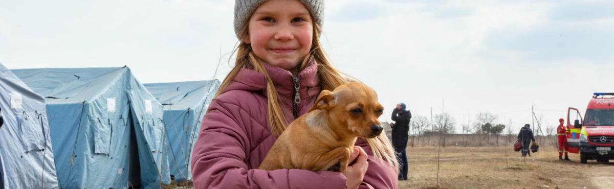 Pipopäinen hymyilevä tyttö seisoo ulkona ja pitää sylissään pientä koiraa. Taustalla näkyy sinisiä telttoja. 