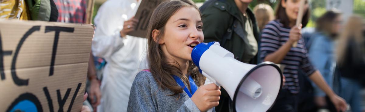 Teini-ikäinen puhuu megafoniin. Taustalla näkyy pahvista tehty mielenosoituskyltti ja muita ihmisiä.