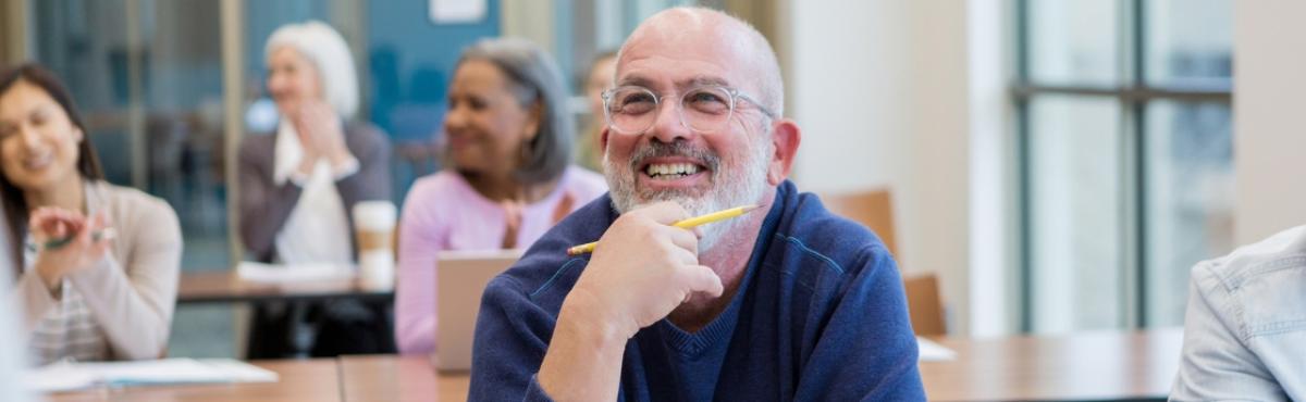 Keski-ikäinen mies silmälasipäinen mies katsoo vasempaan ja hymyilee. Hänellä on kädessään kynä ja hän koskettaa leukaansa kädellään. Taustalla näkyy kolme muuta ihmistä istumassa riveissä olevien pöytien ääressä. 