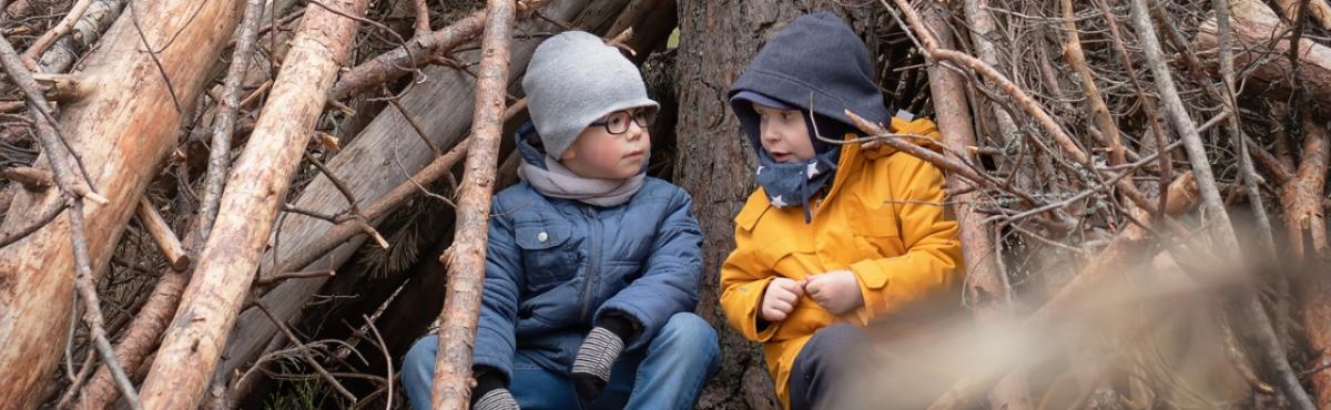 Kaksi välikausivaatteisiin pukeutunutta leikki-ikäistä lasta istuu vierekkäin puumajassa puunrunkoon nojaten. 